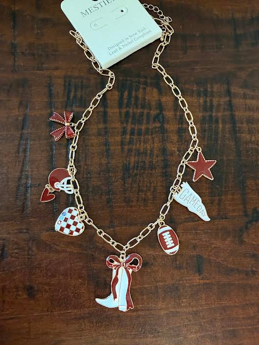 A gold-tone chain necklace with multiple sports-themed charms, including a football, cowboy boots, a star, a heart, and a bow, displayed on a dark wood background with its original white paper tag attached.