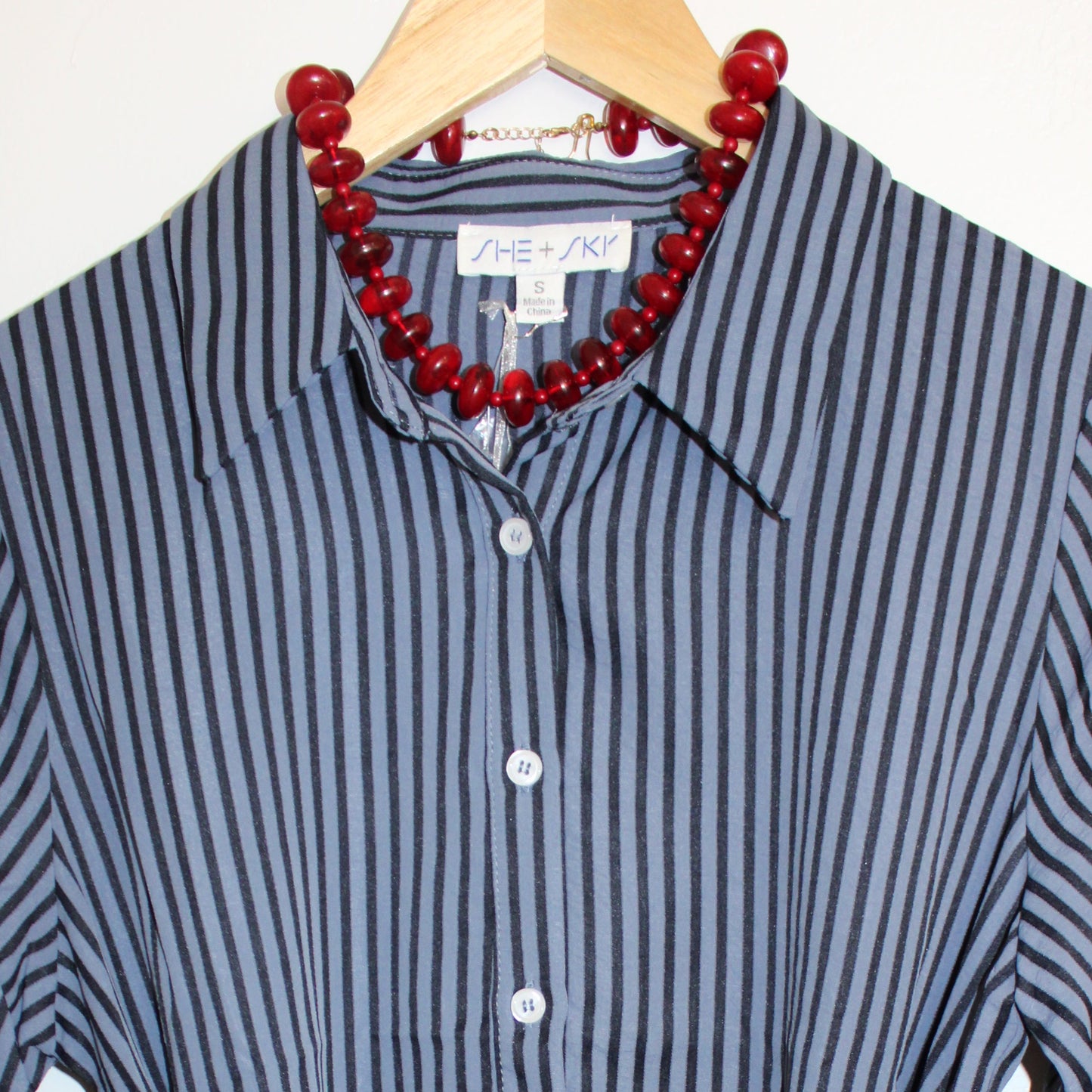 A long-sleeve button-front shirt dress with vertical navy and white stripes, featuring a self-tie waist belt and a red beaded necklace draped over the collar, hanging on a wooden hanger against a white curtain background.