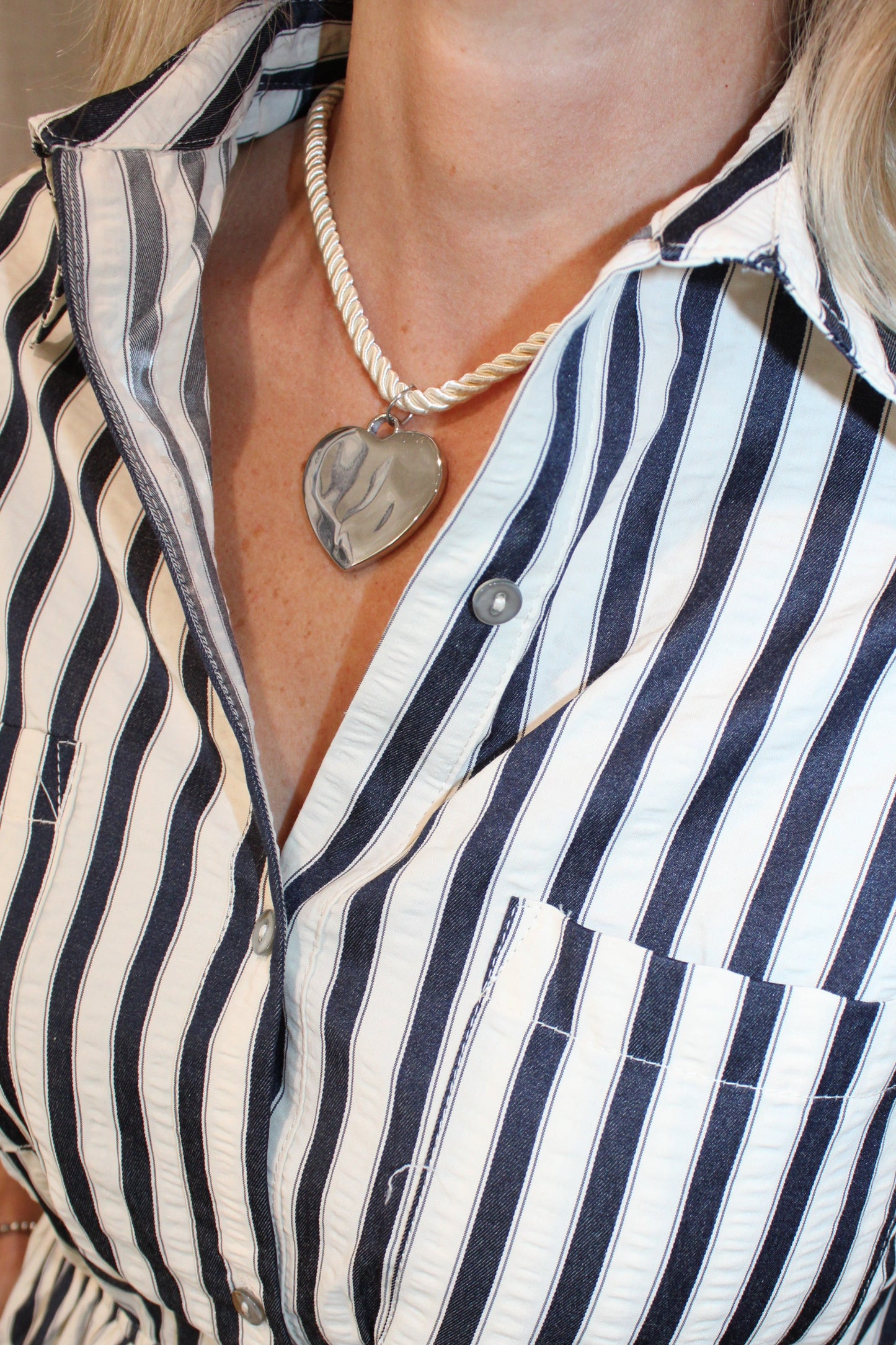 Close-up of a woman wearing a black and white vertically striped shirt dress with a collar, button front, chest pocket, and side tie waist. She is also wearing a rope necklace with a heart-shaped pendant.
