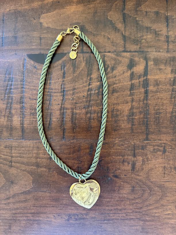 A twisted sage green cord necklace with a gold heart-shaped pendant featuring an engraved profile, displayed on a dark wood background.