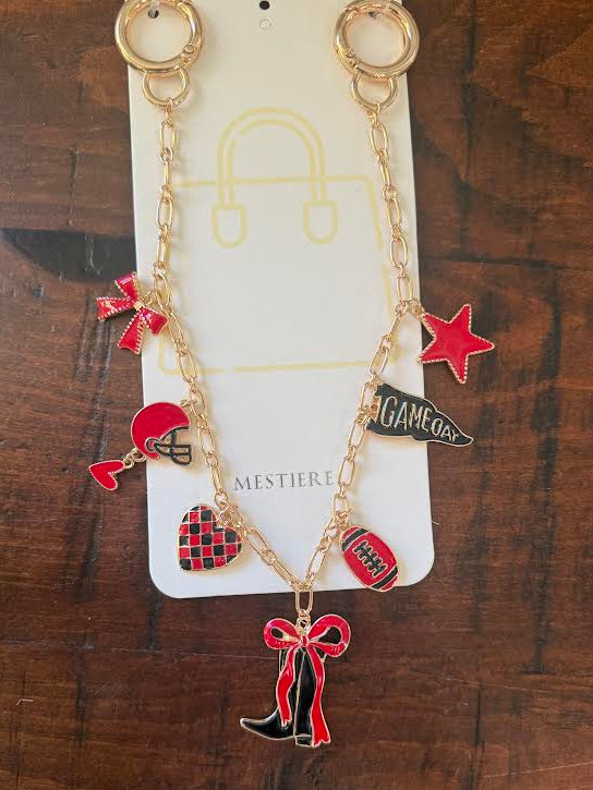 A gold-tone keychain or bag charm necklace with red and black enamel sports-themed charms, displayed on a white card labeled MESTIERE over a dark wood background.