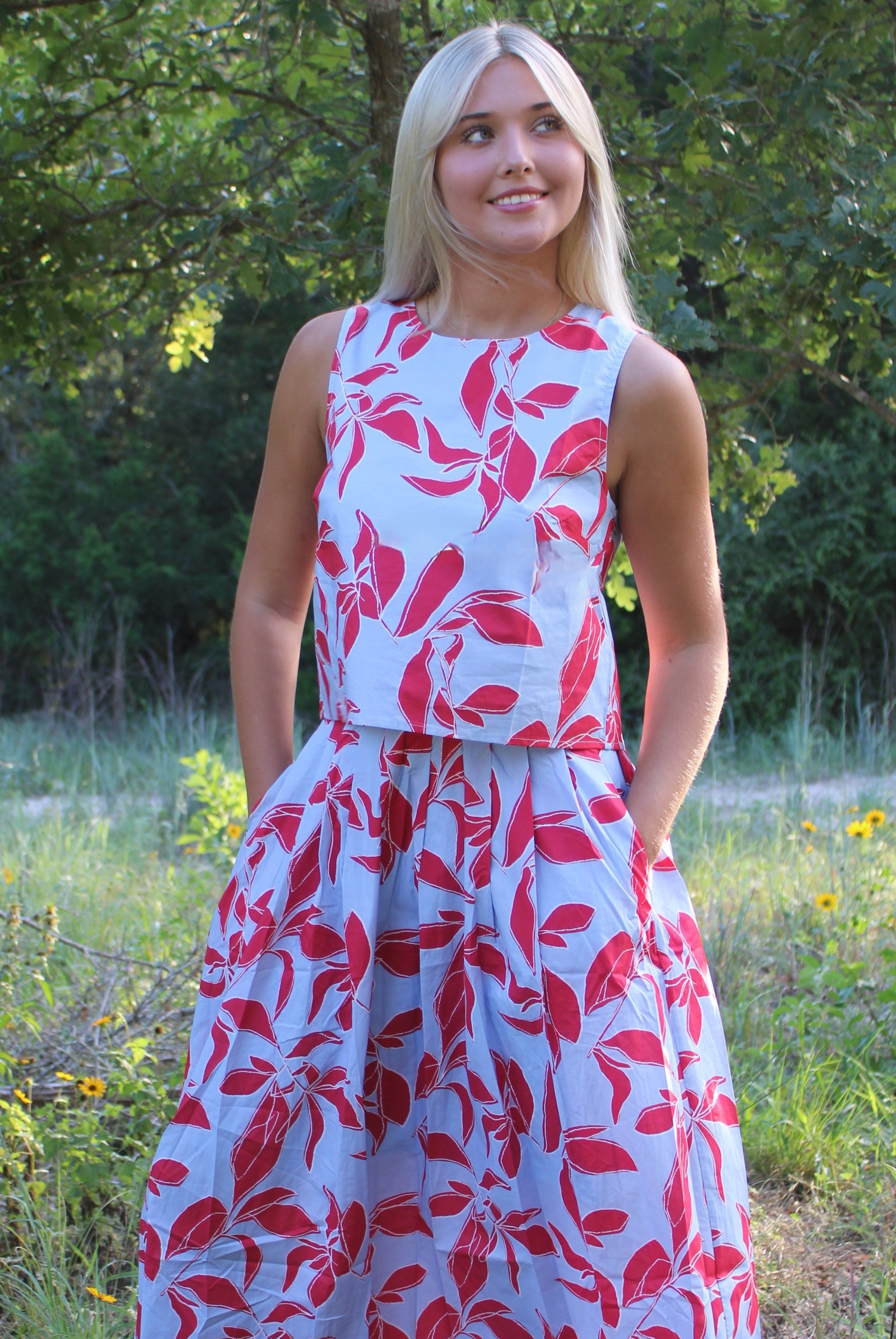 A blonde woman wearing a sleeveless white dress with a red leaf pattern, standing outdoors in a grassy field with trees in the background.