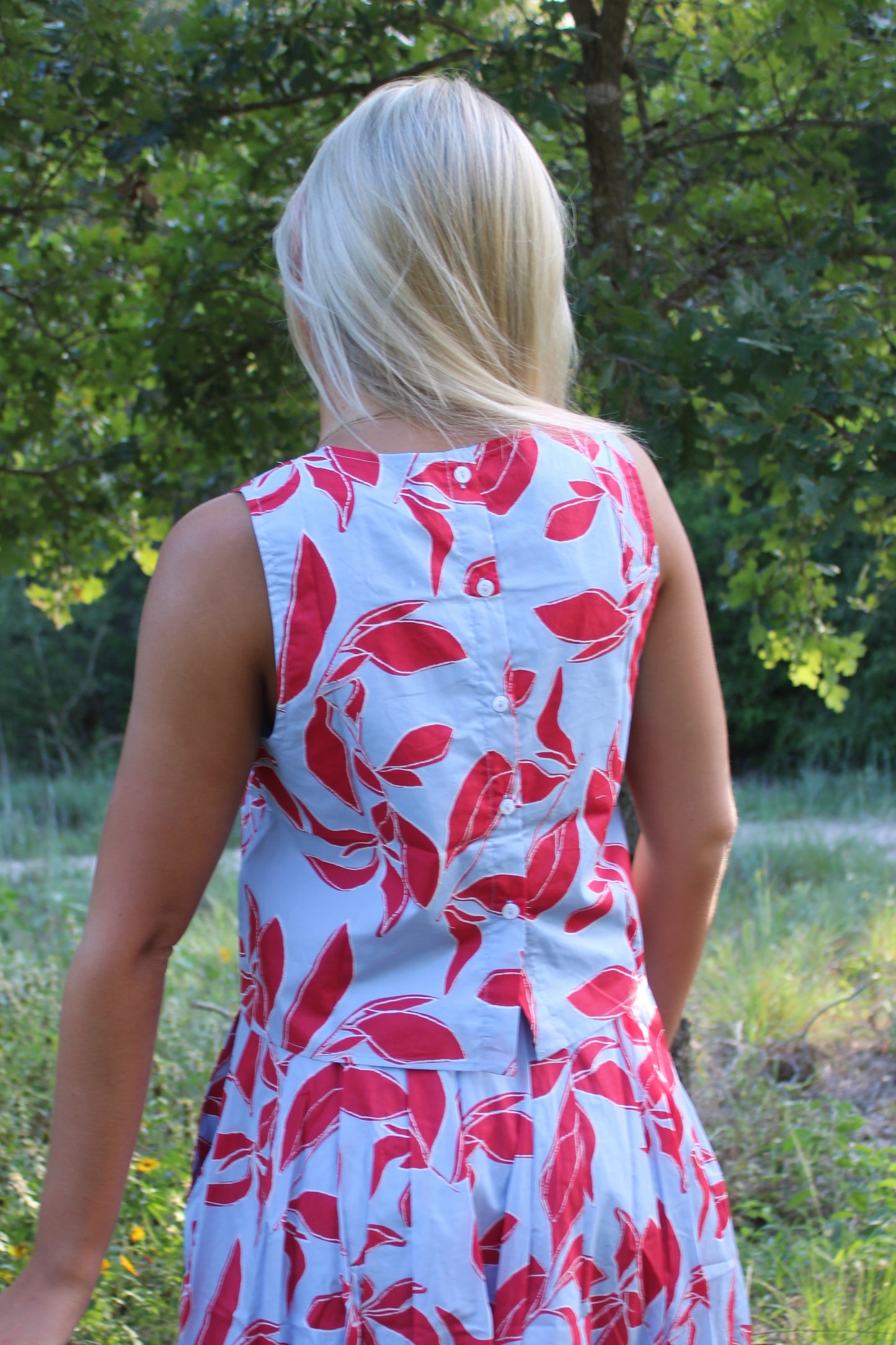 A woman viewed from behind wearing a sleeveless white dress with a red leaf pattern, standing in a grassy outdoor area with trees in the background.