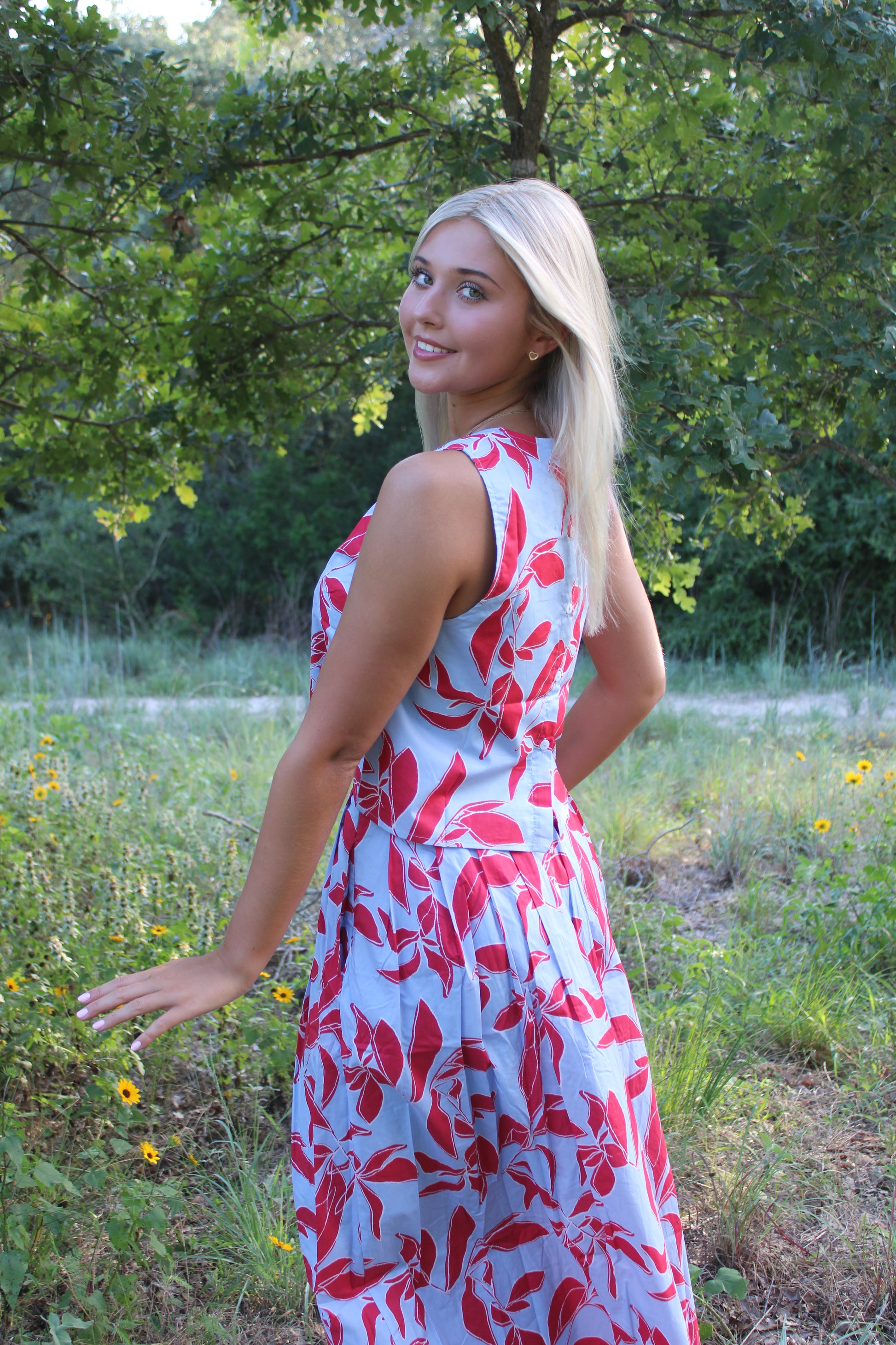 A blonde woman wearing a sleeveless light blue dress with a red floral pattern, standing in a grassy outdoor field with trees in the background.