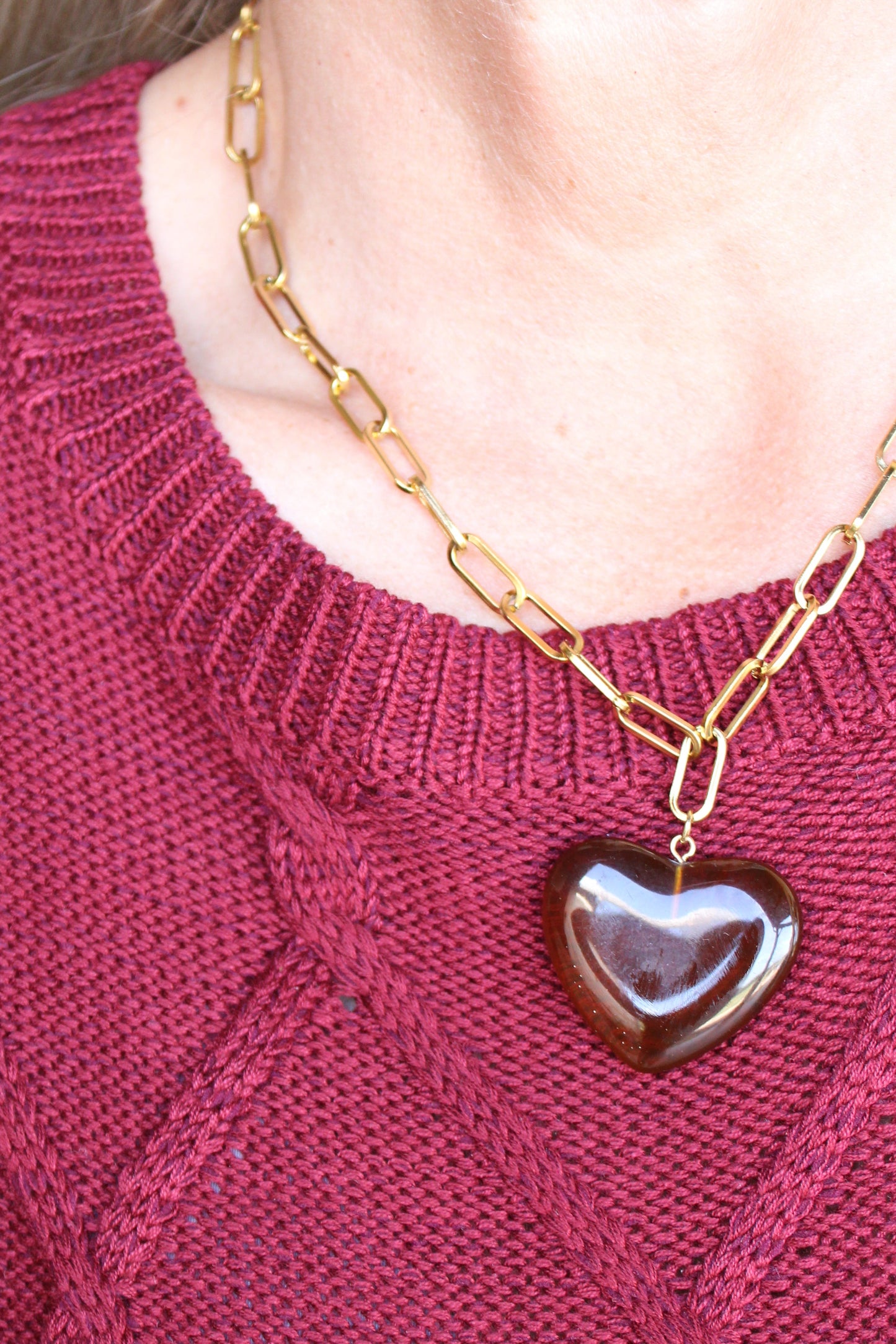 A close-up of a person wearing a burgundy cable-knit sweater and a gold chain necklace with a dark brown heart-shaped pendant.