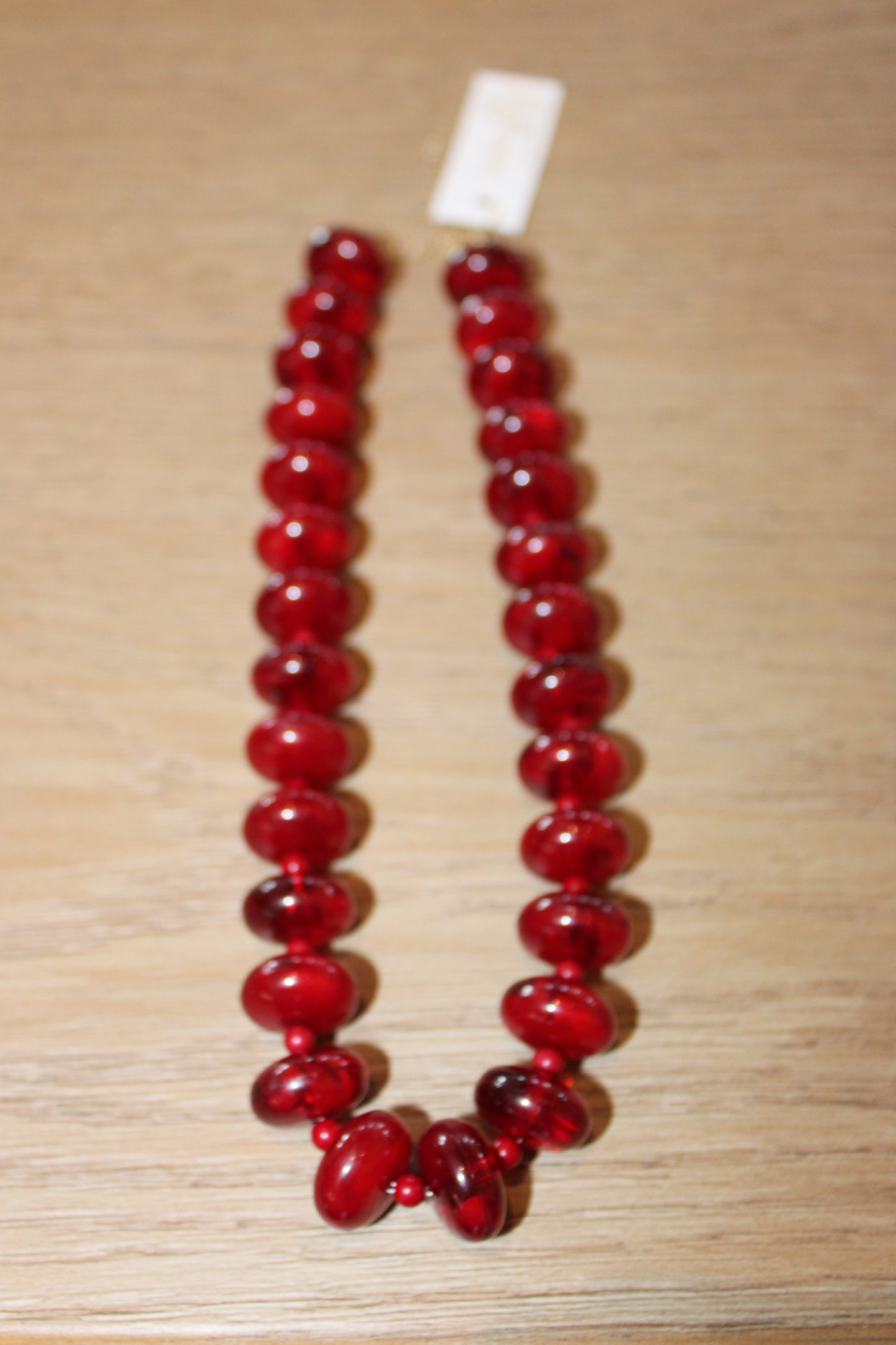 A red beaded necklace with glossy, polished oval beads and a small white tag at the top, displayed on a light wood surface.