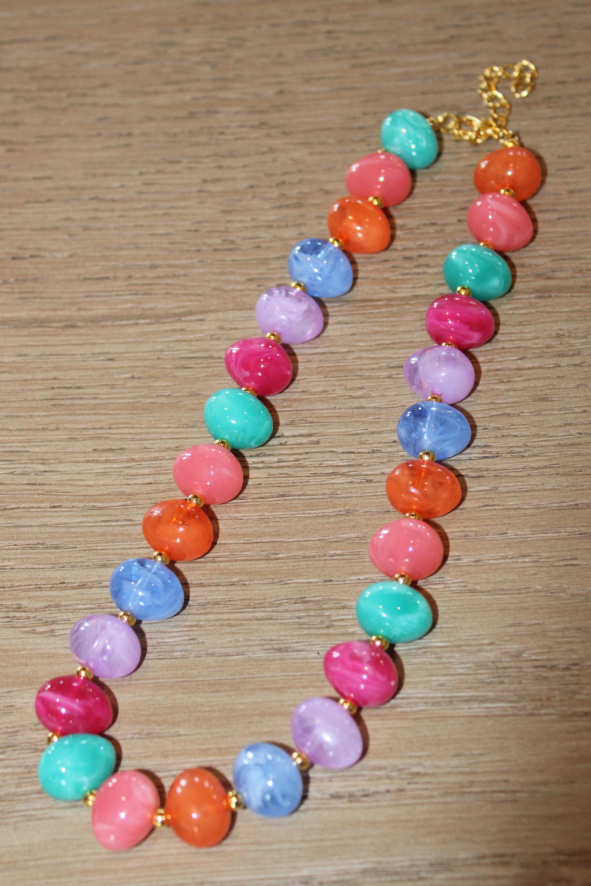 A colorful beaded necklace with round, marbled beads in pink, orange, teal, blue, purple, and red, separated by small gold spacers, displayed on a wooden surface.