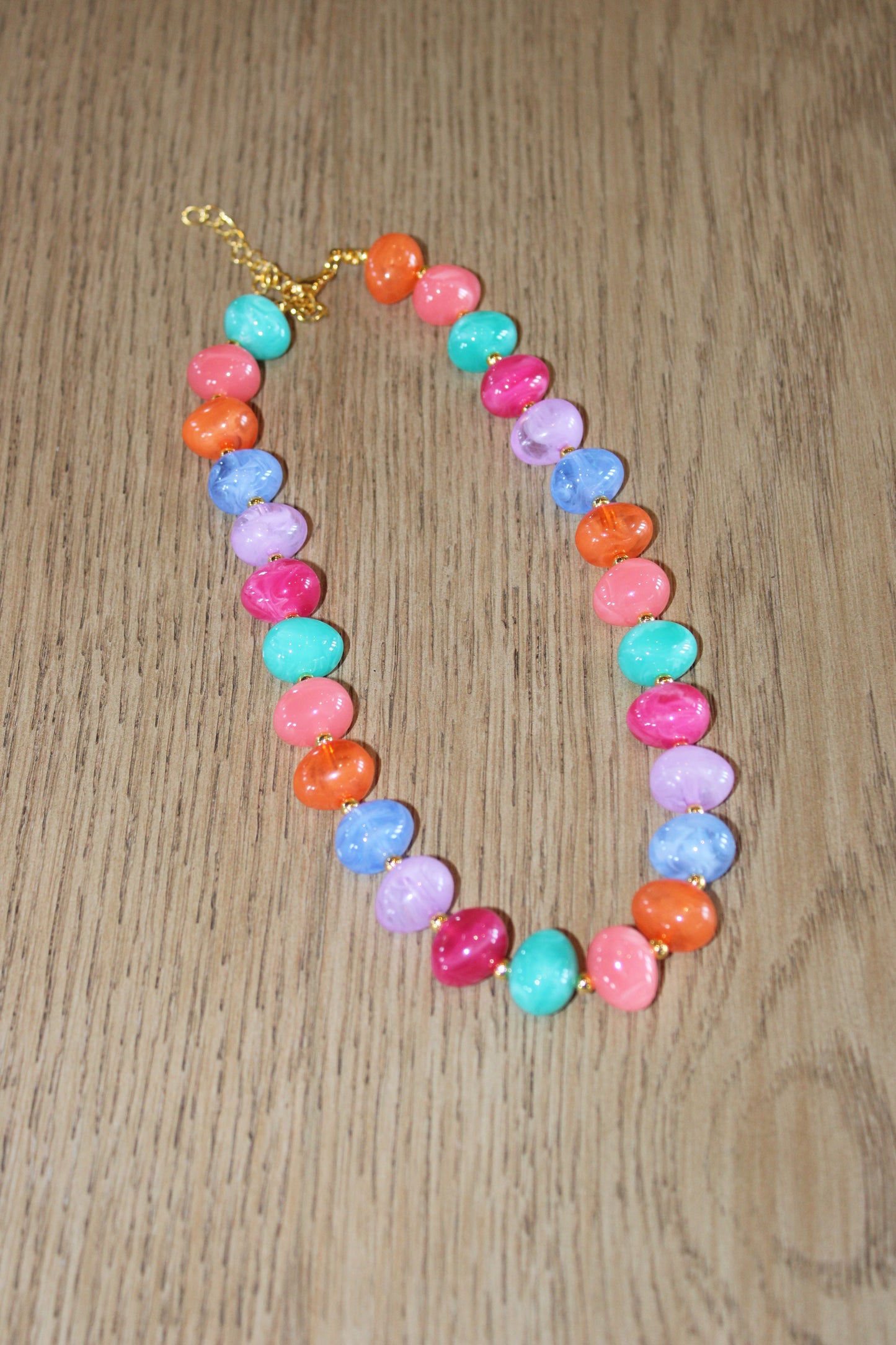 A colorful beaded necklace with marbled pastel and bright beads in pink, orange, turquoise, purple, and blue, arranged on a light brown wooden surface.