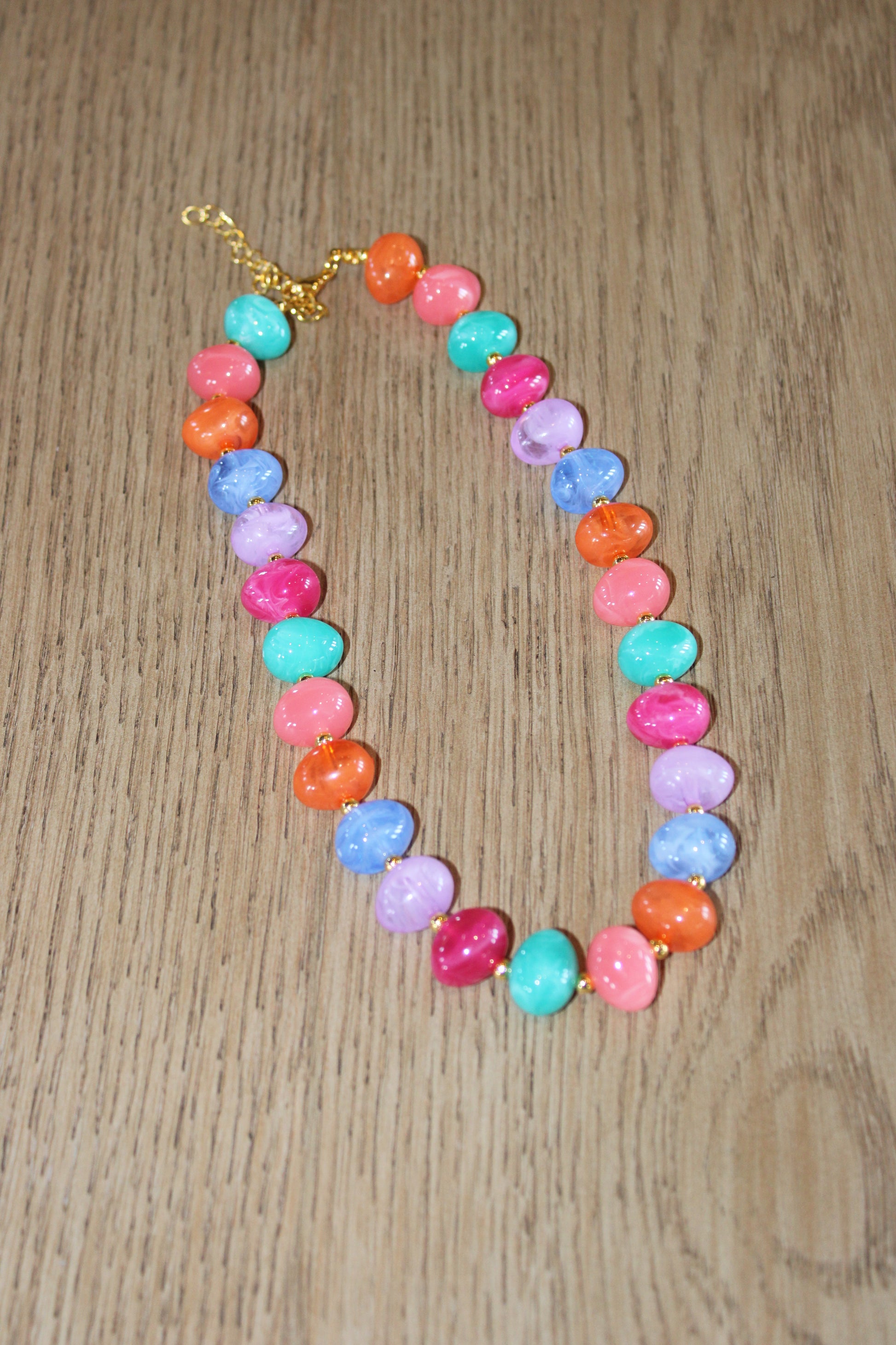 A colorful beaded necklace with marbled pastel and bright beads in pink, orange, turquoise, purple, and blue, arranged on a light brown wooden surface.