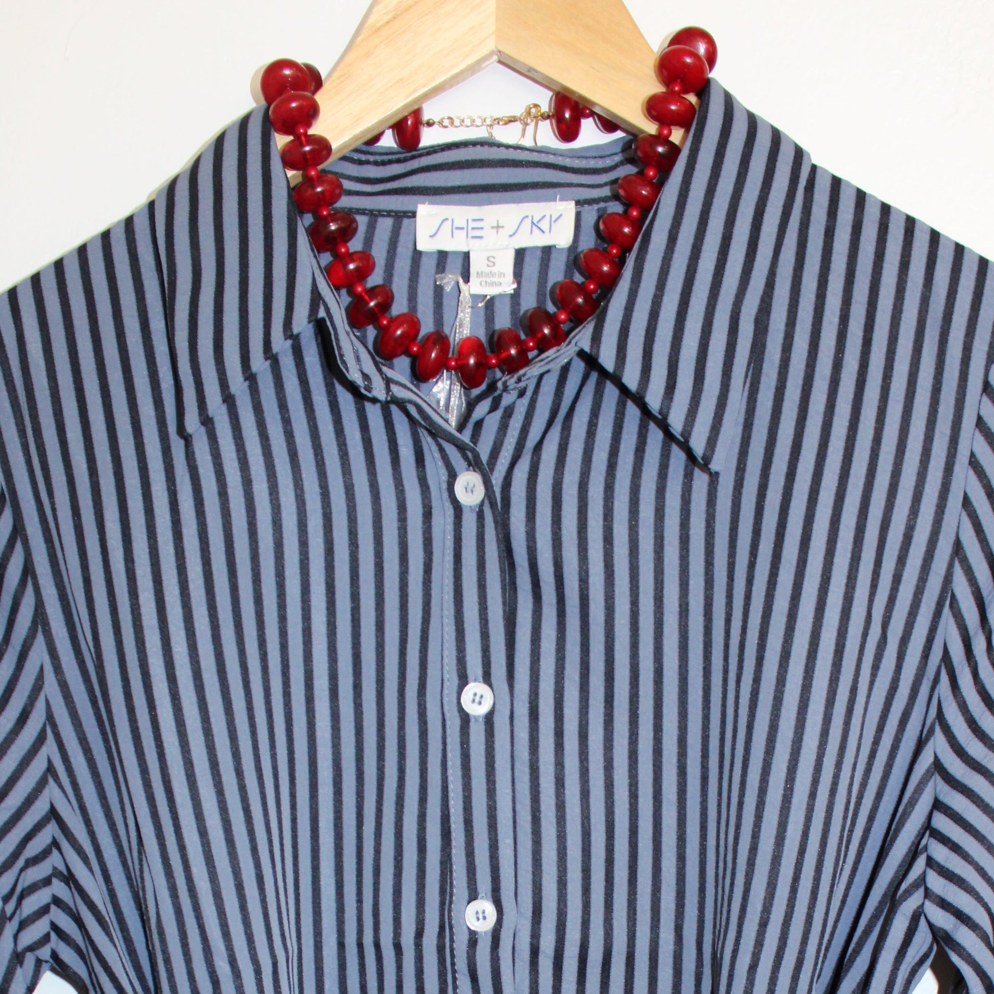 A long-sleeve button-front shirt dress with vertical navy and white stripes, featuring a self-tie waist belt and a red beaded necklace draped over the collar, hanging on a wooden hanger against a white curtain background.