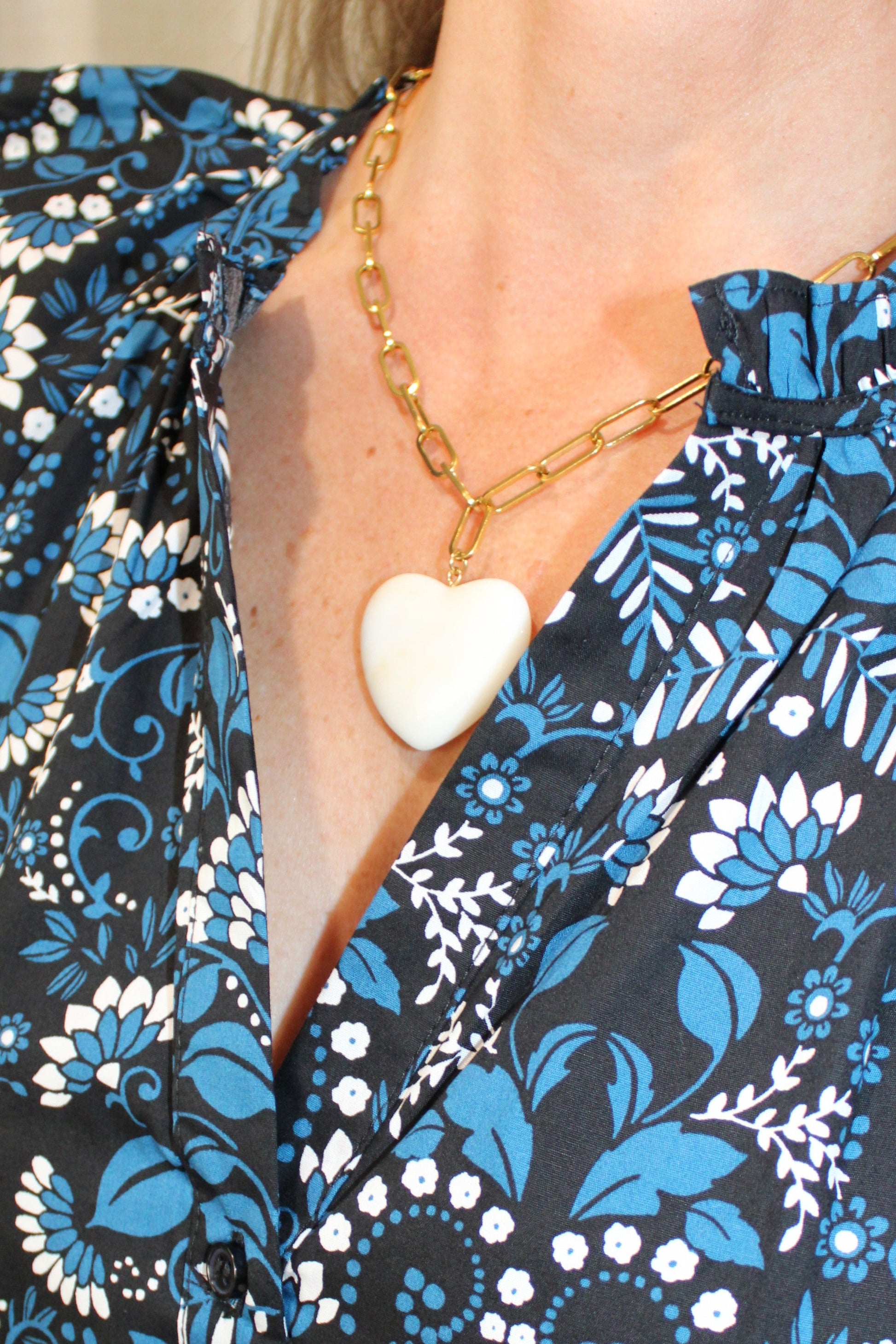A close-up of a woman wearing a black wrap-style blouse with a blue and white floral pattern, styled with a gold chain necklace featuring a white heart-shaped pendant.