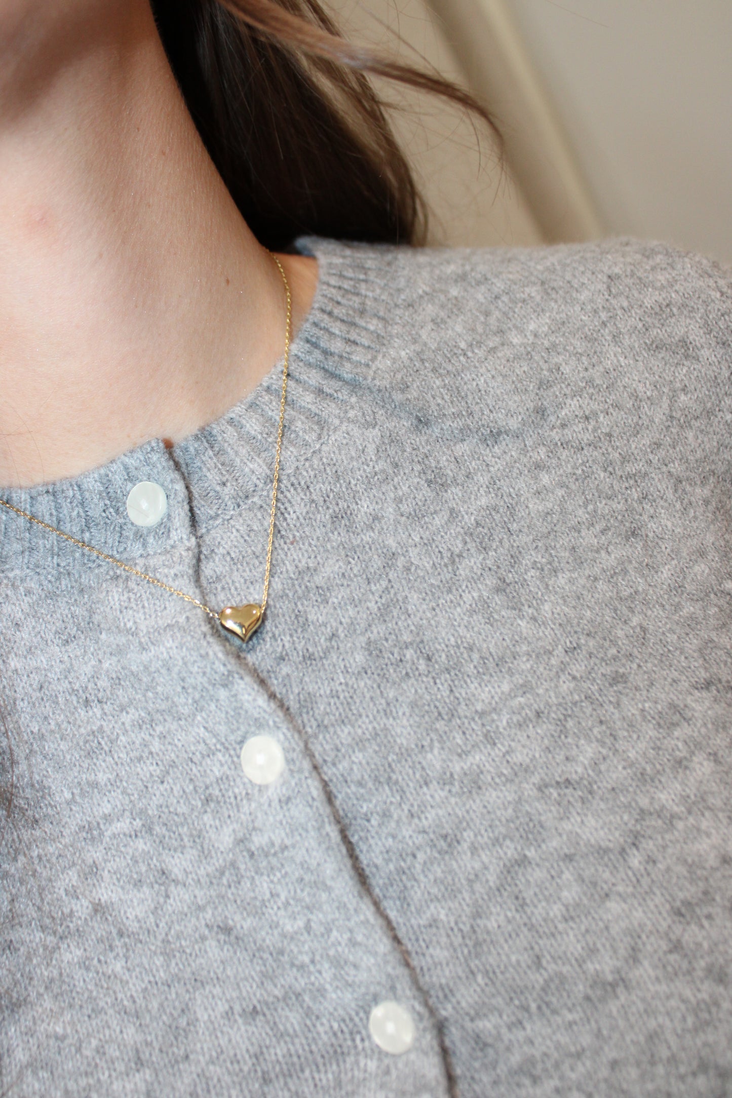 A close-up of a person wearing a delicate gold necklace with a small triangular pendant, styled over a light gray knit cardigan.