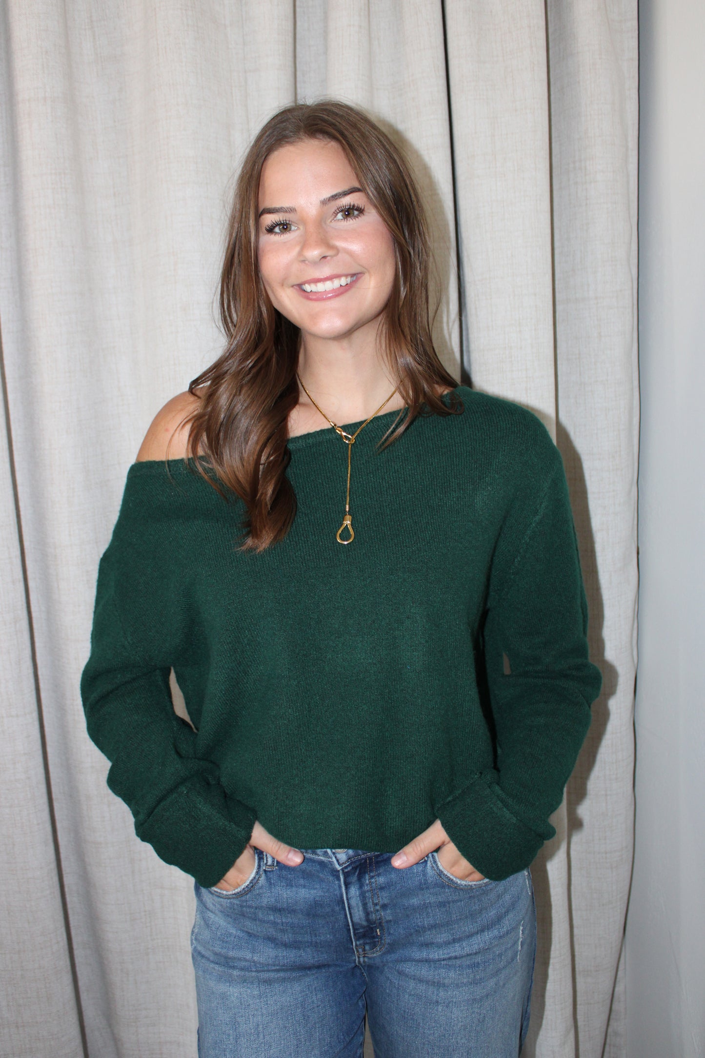 A woman with long brown hair smiling, wearing a dark green off-the-shoulder knit sweater and blue jeans, standing in front of a light beige curtain.