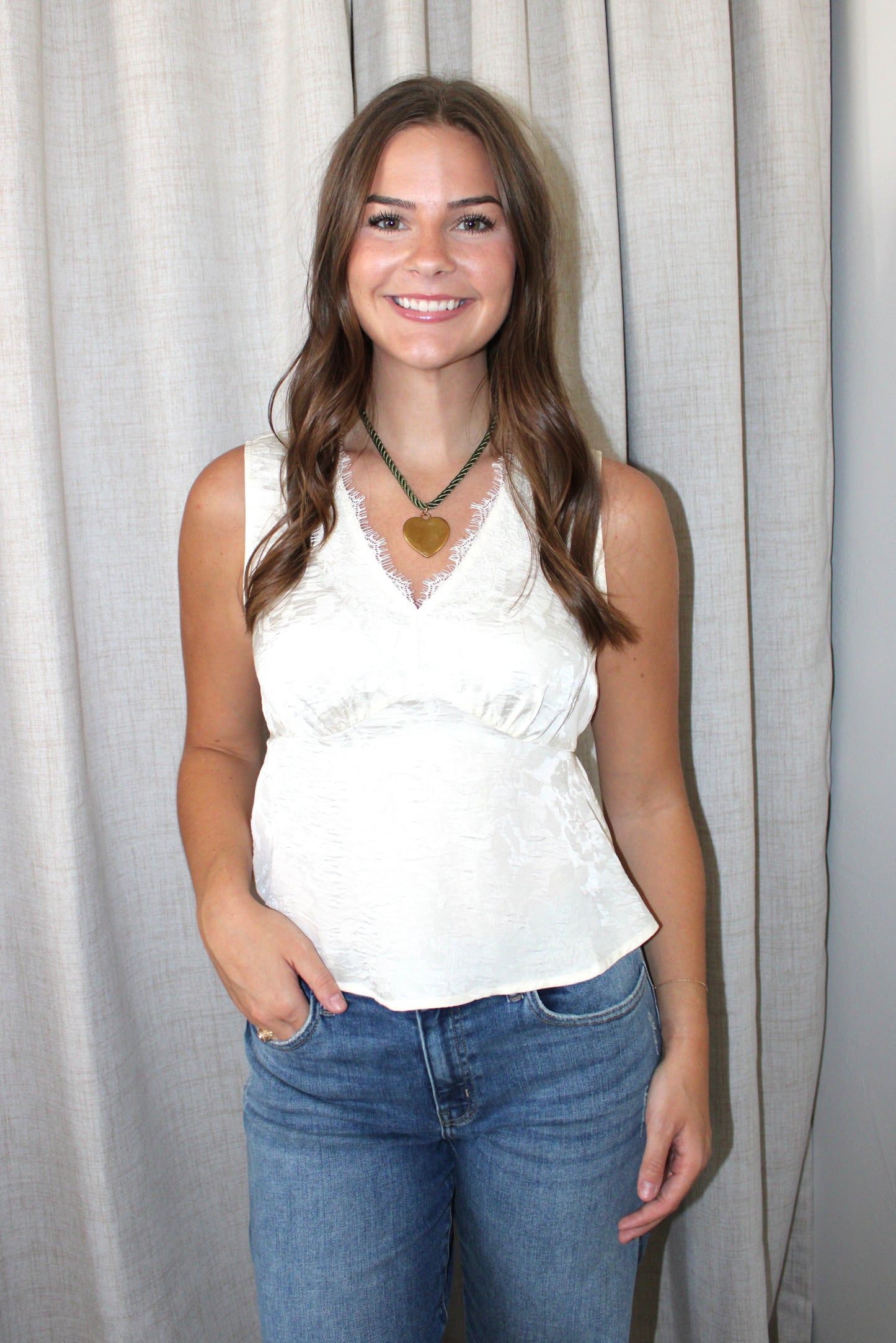 A woman with long brown hair stands in front of a light gray curtain, wearing a white lace-trimmed sleeveless top and blue jeans, with a green necklace featuring a gold heart pendant.
