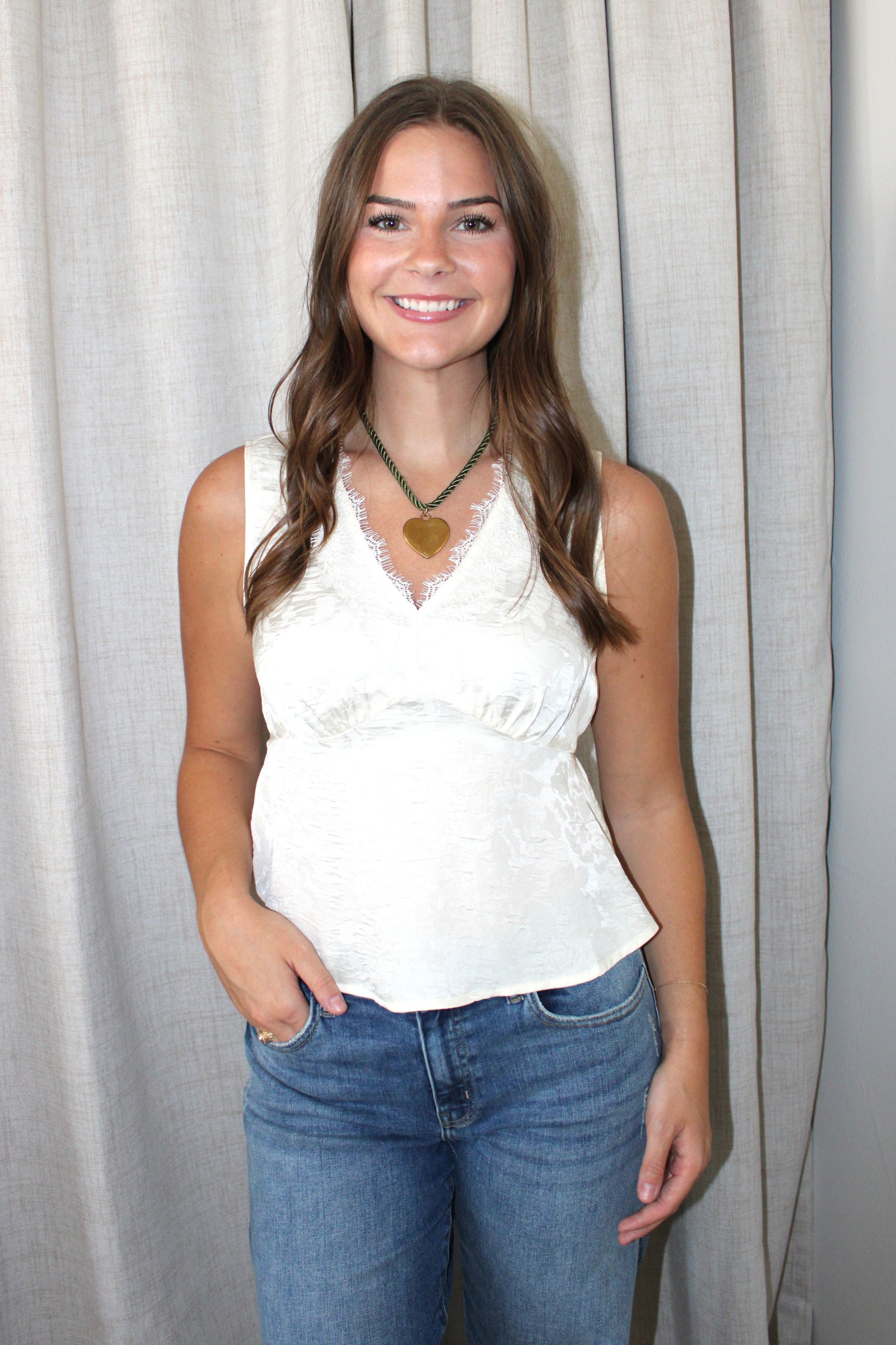A woman with long brown hair stands in front of a light gray curtain, wearing a white lace-trimmed sleeveless top and blue jeans, with a green necklace featuring a gold heart pendant.