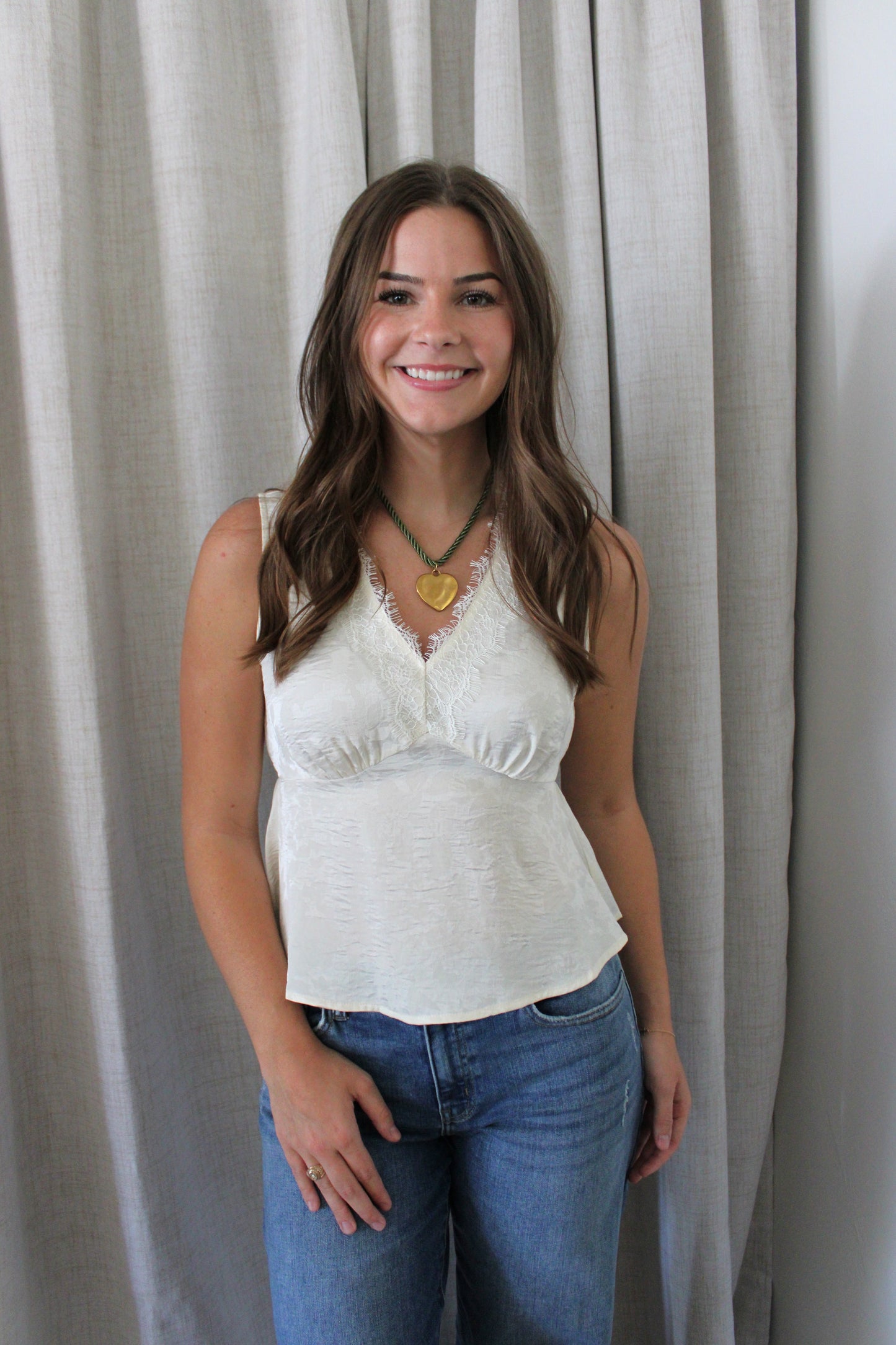A woman with long brown hair stands in front of light gray curtains, wearing a white lace-trimmed camisole top and blue jeans.