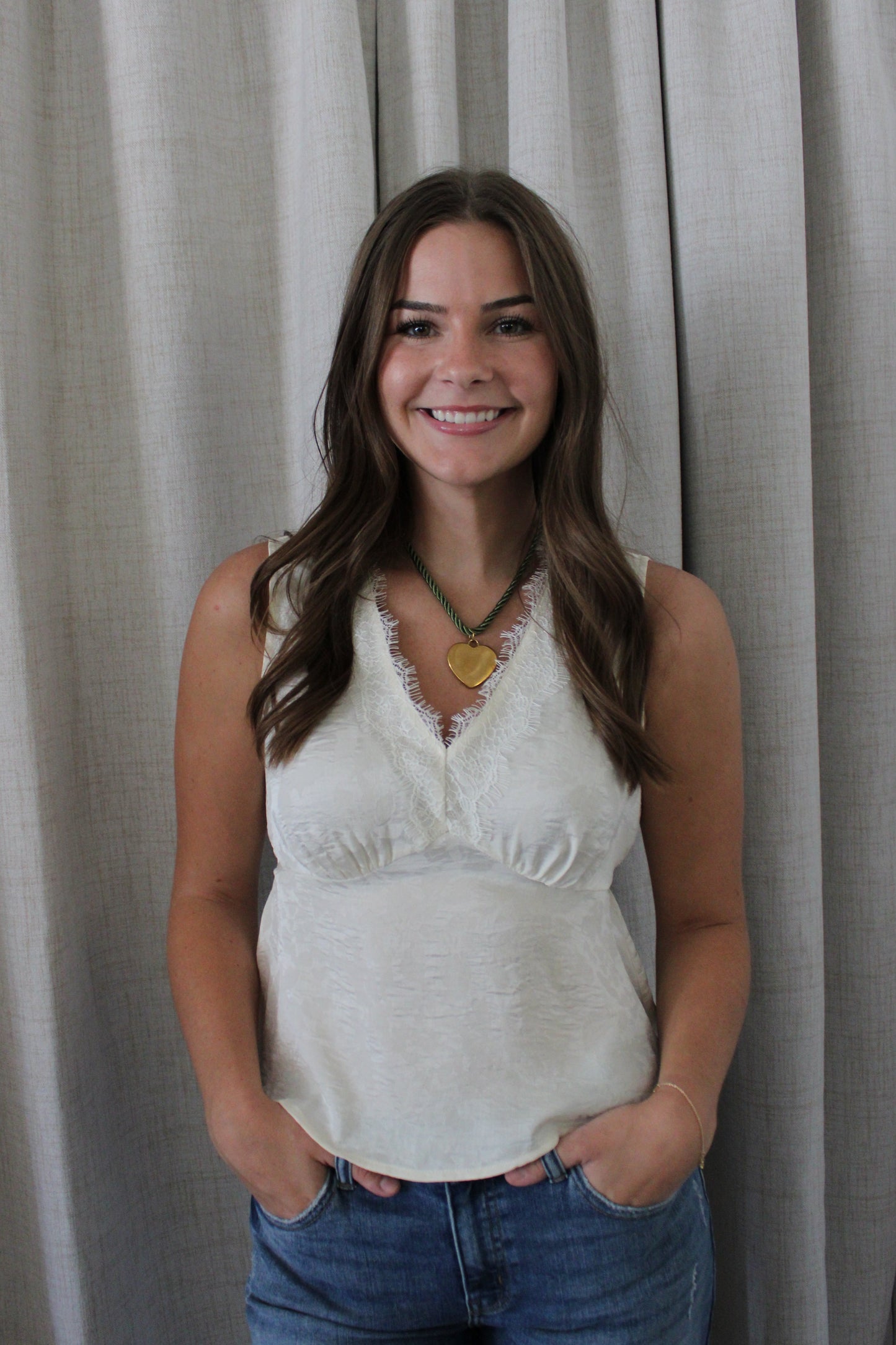A smiling woman with long brown hair stands in front of light gray curtains, wearing a sleeveless cream-colored lace-trimmed top and blue jeans.