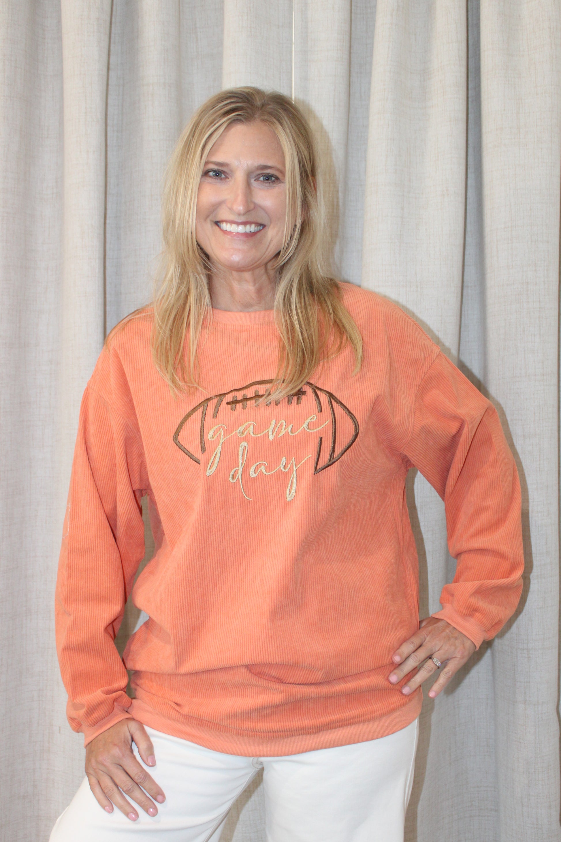 A smiling blonde woman wearing a peach-orange long-sleeve sweatshirt with a football graphic and the words 'game day' in script, paired with white pants, standing in front of a light beige curtain backdrop.